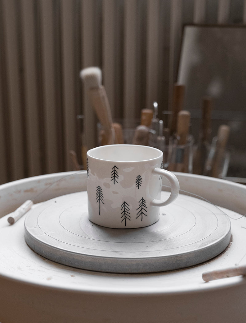 A white porcelain cup with a pine tree motif pattern shown on a throwing wheel with tools in the background.