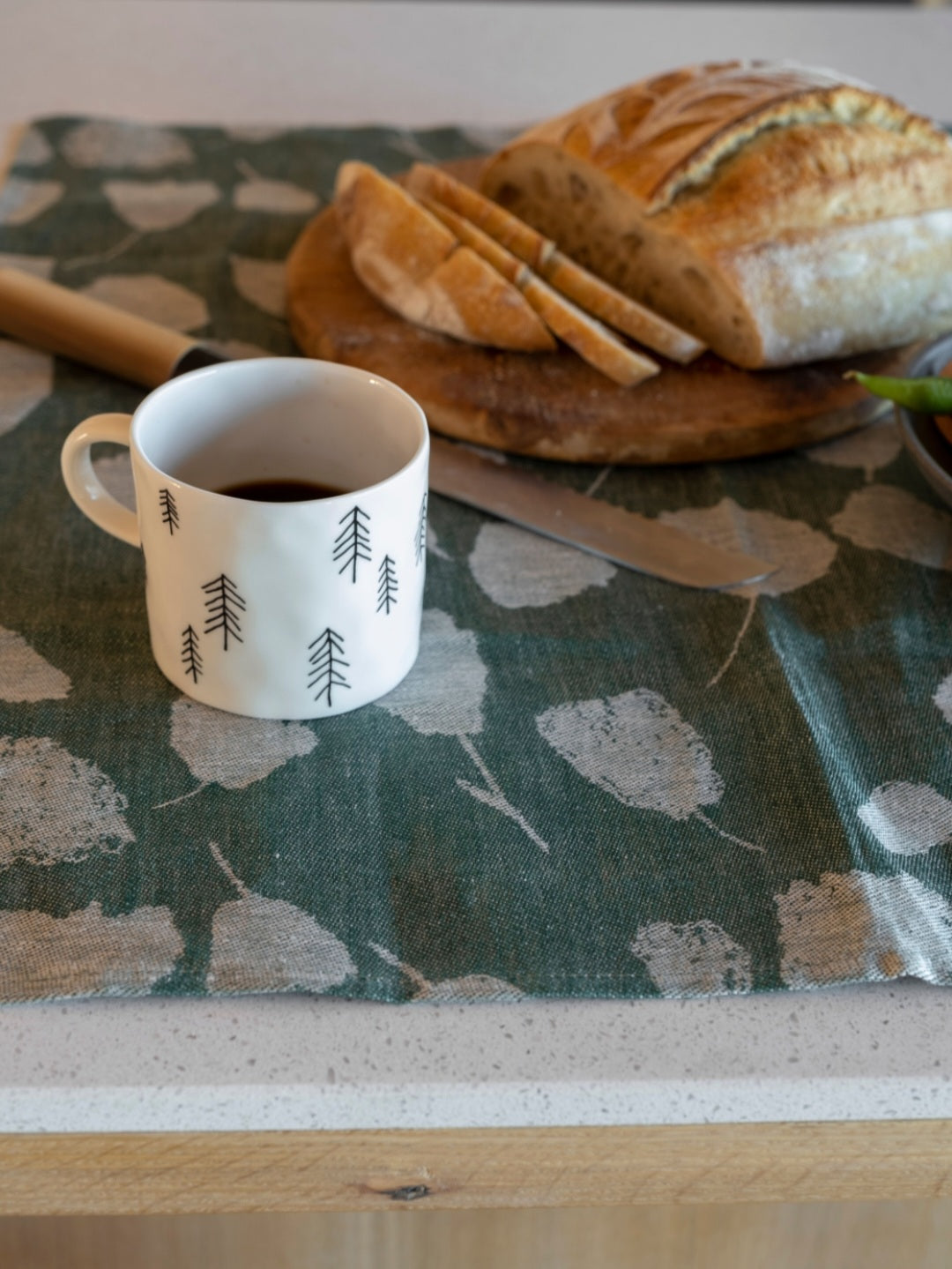 A white porcelain cup with a pine tree motif pattern placed on green bouquet tea towel with a piece of bread and a knife in the background.