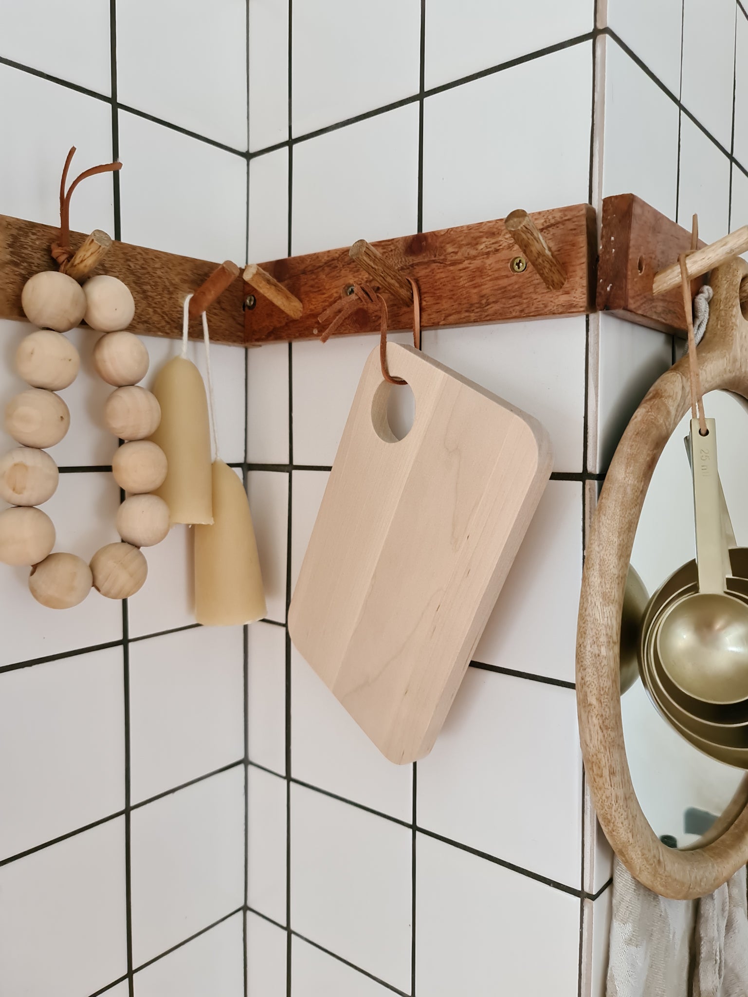 A small birch wood serving board with a hole in the corner, hanging on a wall with other wooden kitchen items.