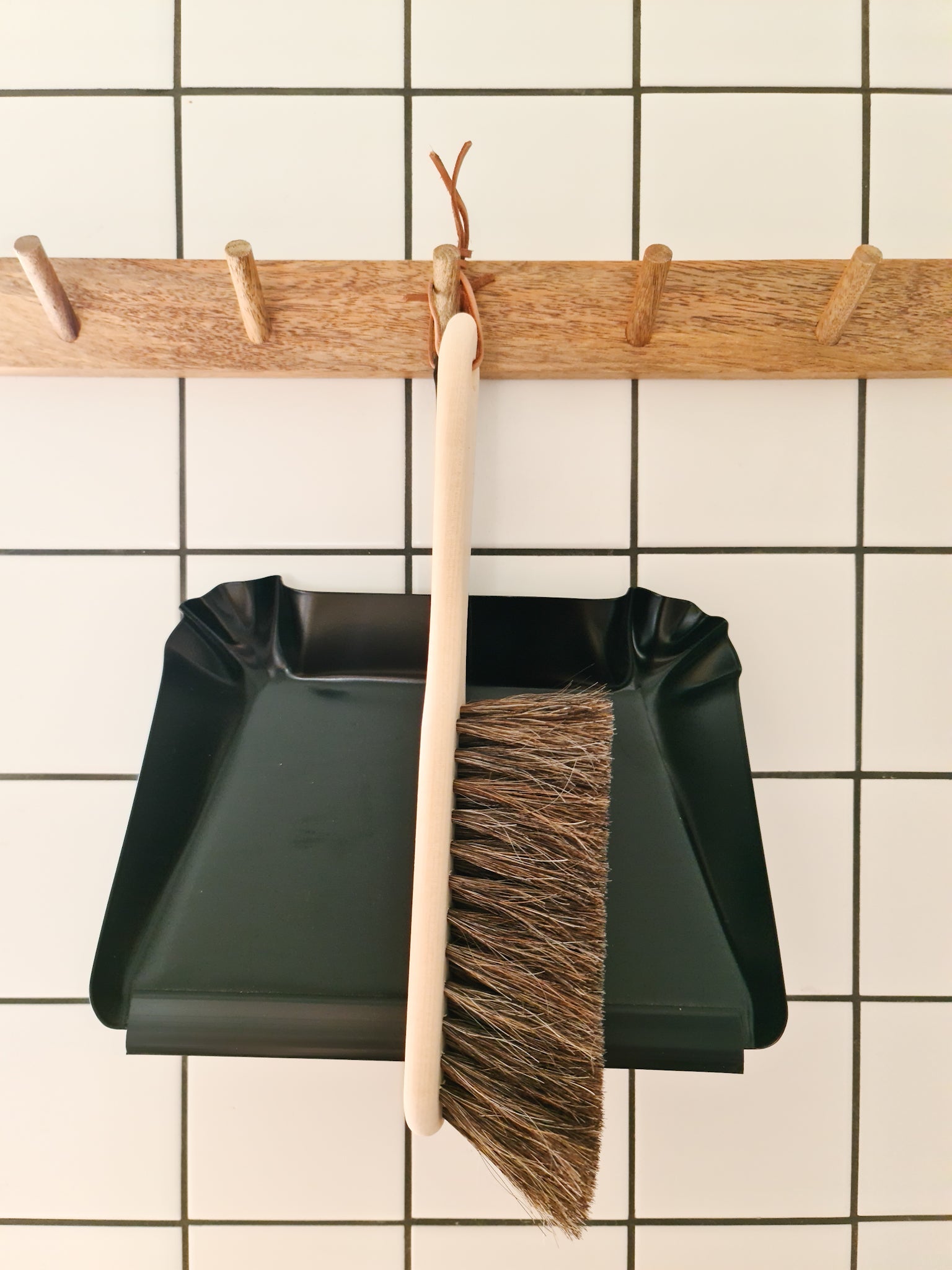 A broom made of oil treated Beech wood with Horse Hair hanging off a wooden peg displayed alongside a black metal dust pan against a tiled background.