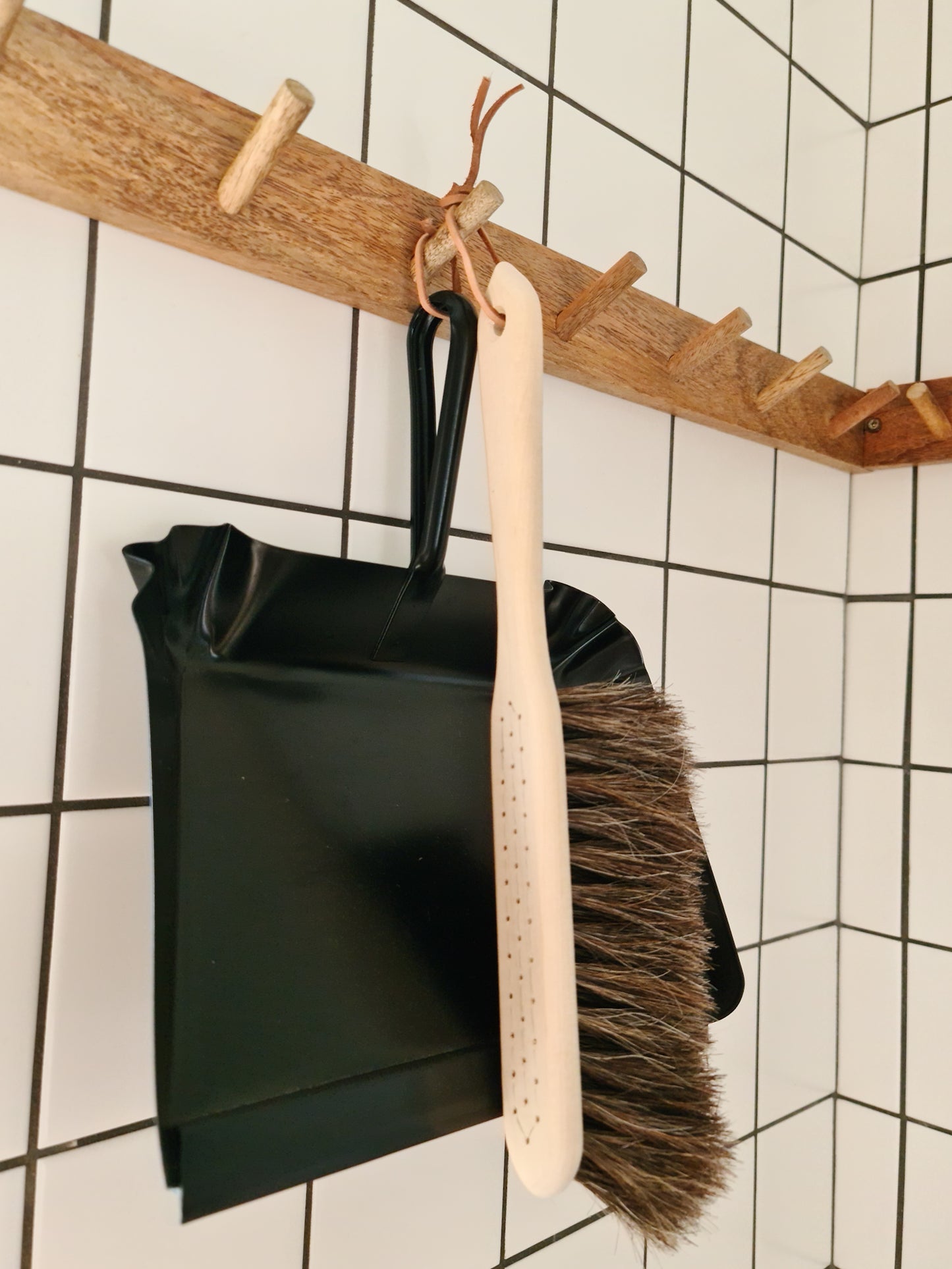 A broom made of oil treated Beech wood with Horse Hair hanging off a wooden peg displayed alongside a black metal dust pan against a tiled background.