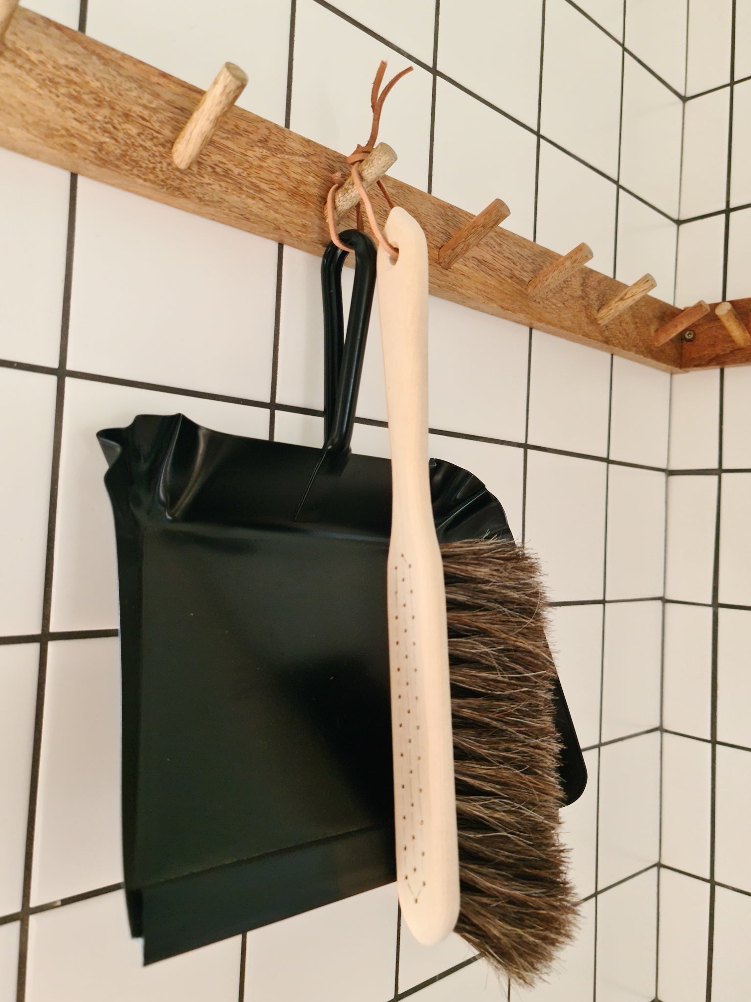 A broom made of oil treated Beech wood with Horse Hair hanging off a wooden peg displayed alongside a black metal dust pan against a tiled background.