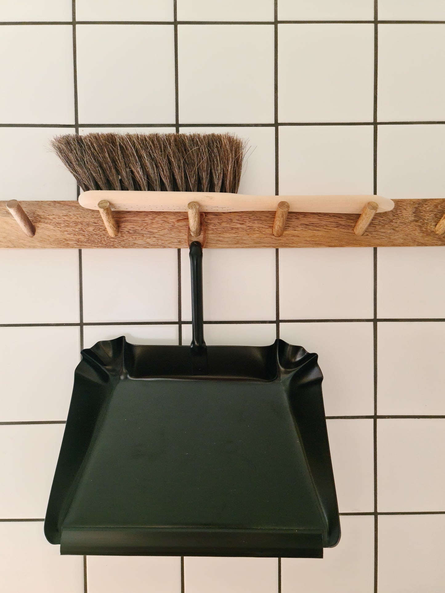 A broom made of oil treated Beech wood with Horse Hair hanging off a wooden peg displayed alongside a black metal dust pan against a tiled background.