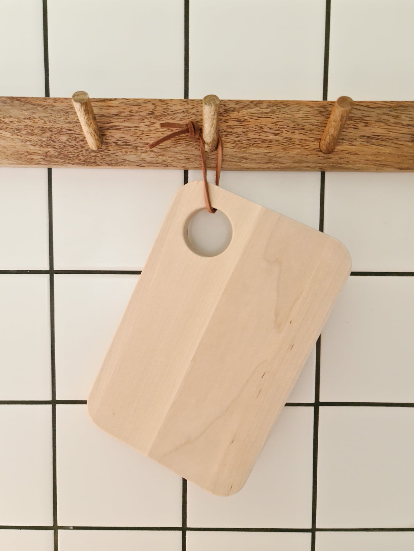 Birch sandwich platter hanging off a wooden peg rail by a leather cord through its hole against a tiled background.
