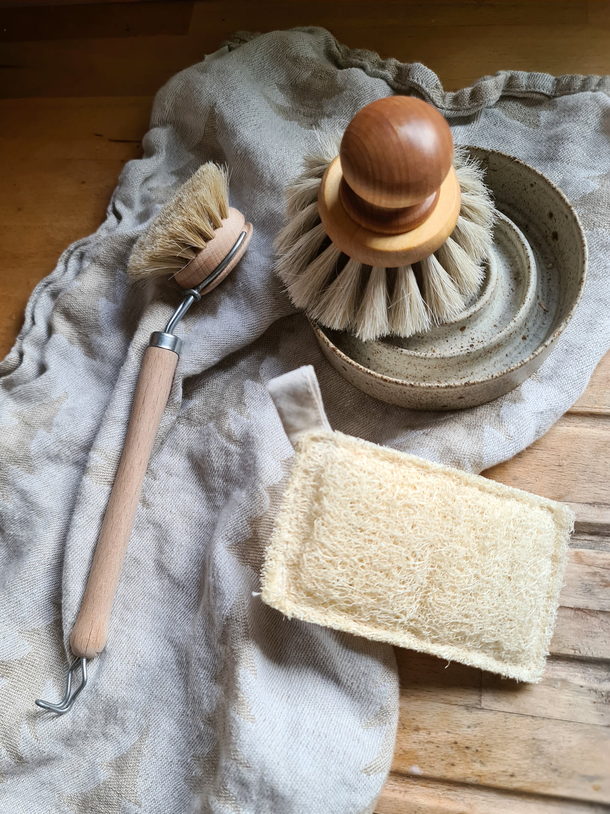 Dish brush laying on a teatowel on a wooden table amongst other kitchen tools.