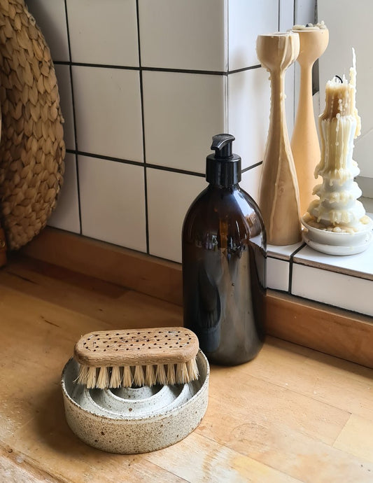 An oak wood nail brush with natural bristles resting on a candle holder next to a glass bottle and candles in the background.