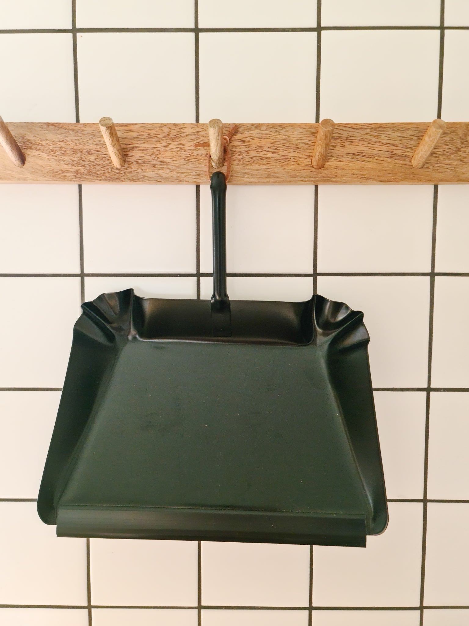 A black metal dustpan with a short handle is hanging on a white tile wall.