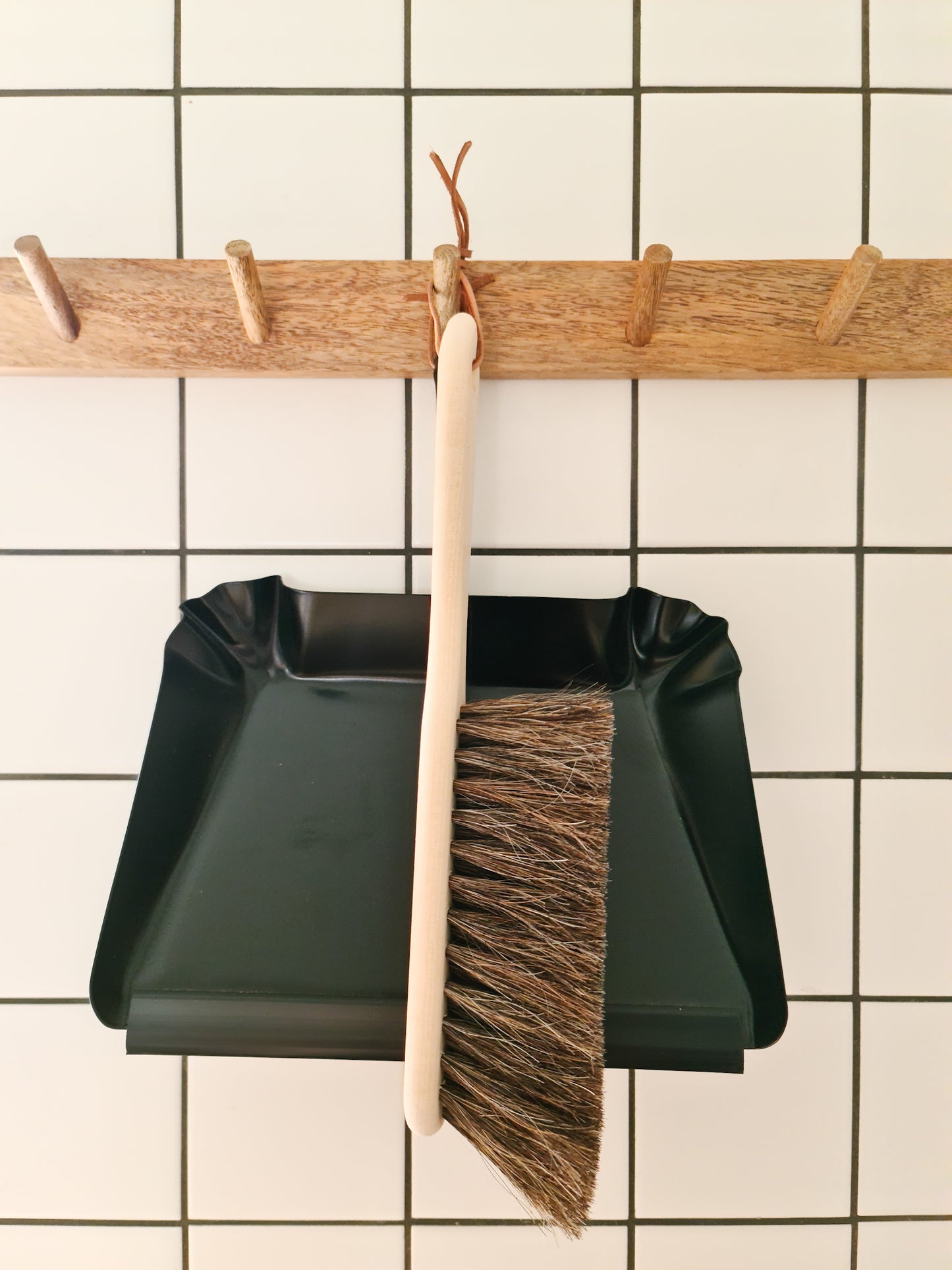 A black metal dustpan with a short handle is hanging on a white tile wall next to a broom with a wooden handle. The broom has natural bristles.