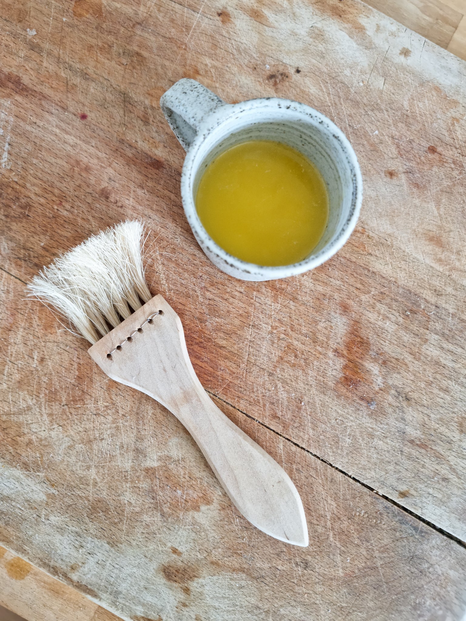 A pastry brush made of birch wood and horse hair, lying next to a small bowl filled with egg wash on a wooden surface.