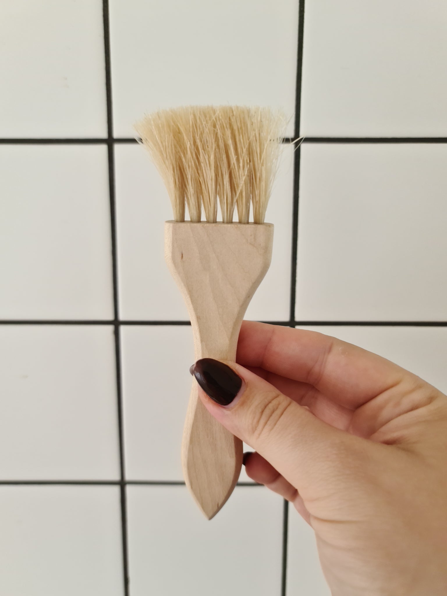 A woman with painted nails holding up a pastry brush made of birch and horsehair against a tiled background.