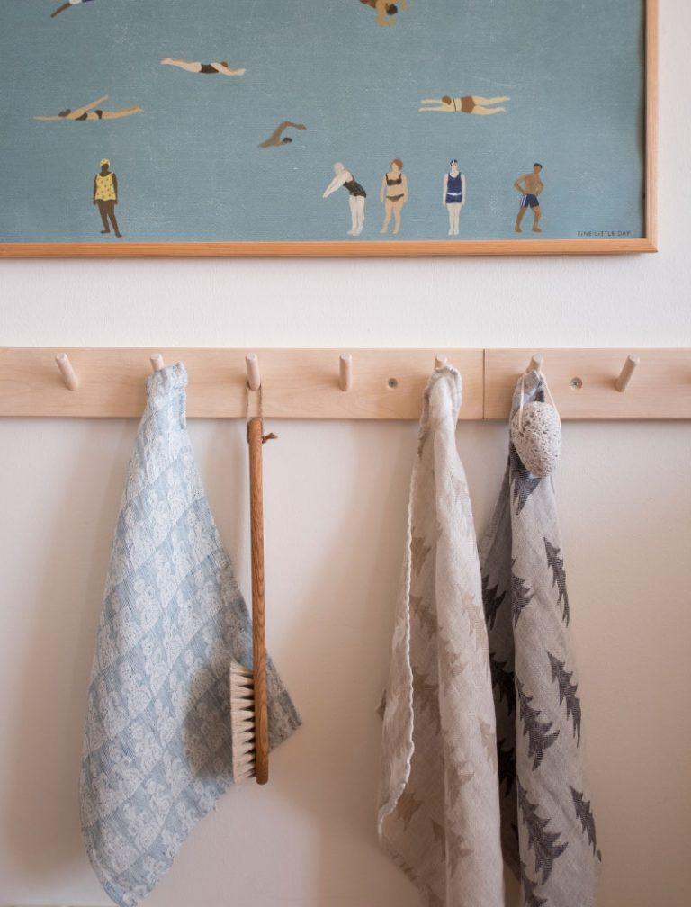 A peg rail rack made of birch wood with multiple hooks, mounted on a wall and holding three tea towels and a brush, beneath a framed print.
