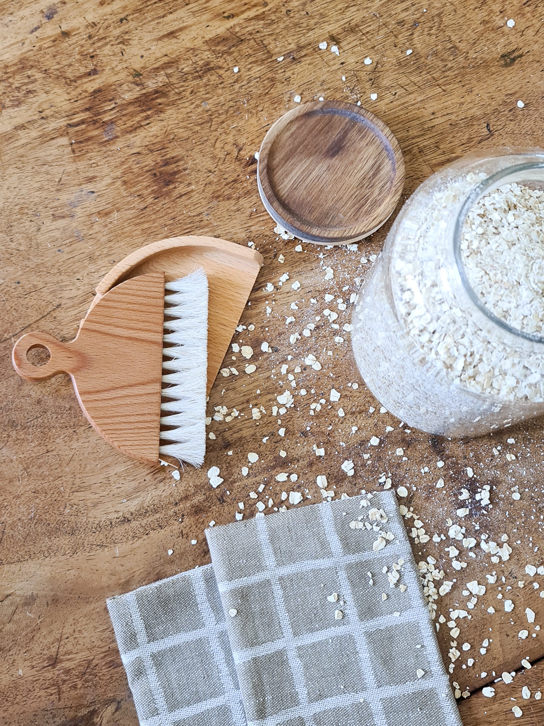 A wooden table brush with a matching dustpan, placed on a wooden surface with a checkered patterned cloth and some scattered crumbs.