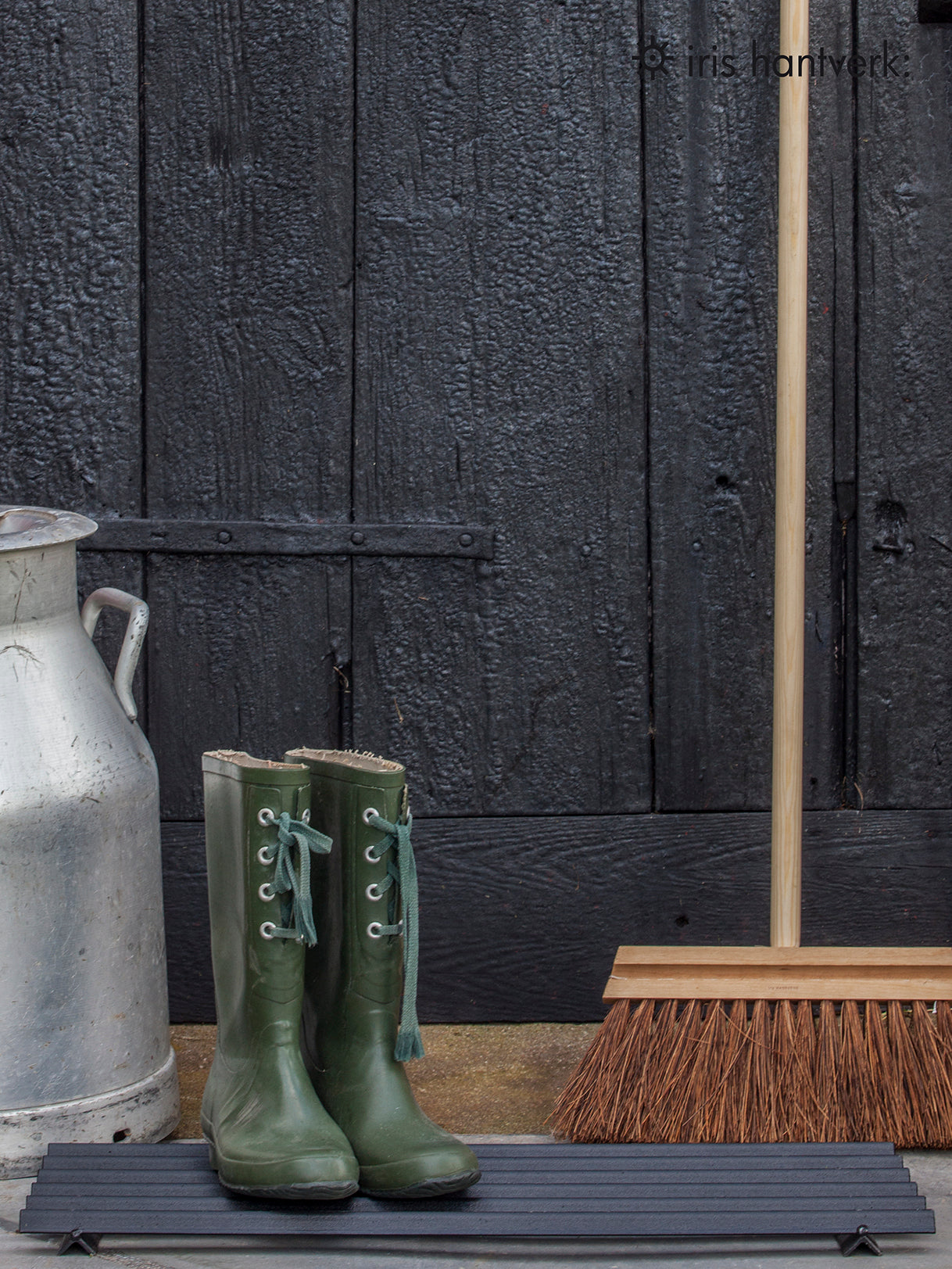 Wooden Broom resting against black wood, with green wellies on a mat placed next to it.