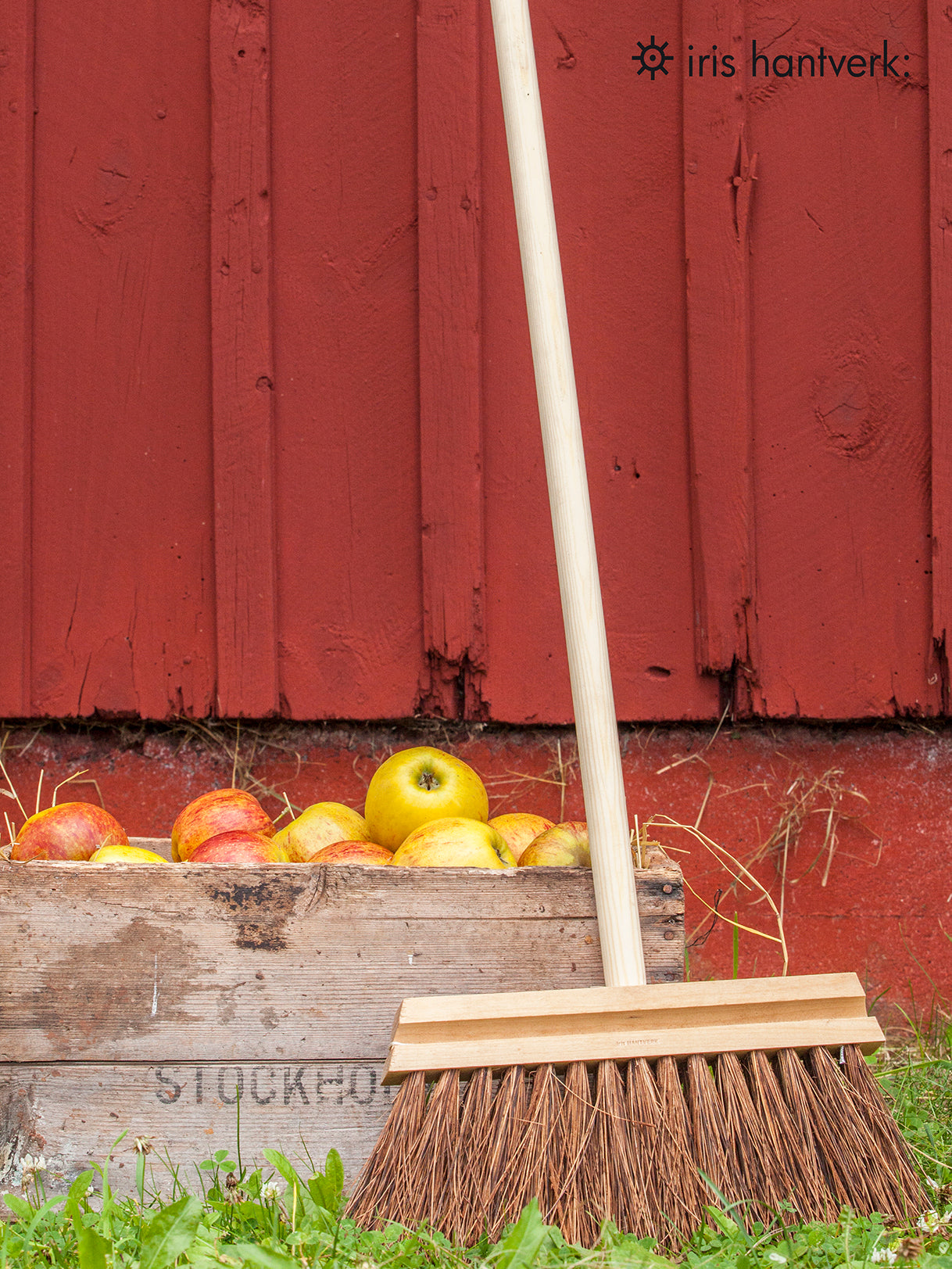 Wooden broom resting against a rate of apples with a terracotta red shed in the background.