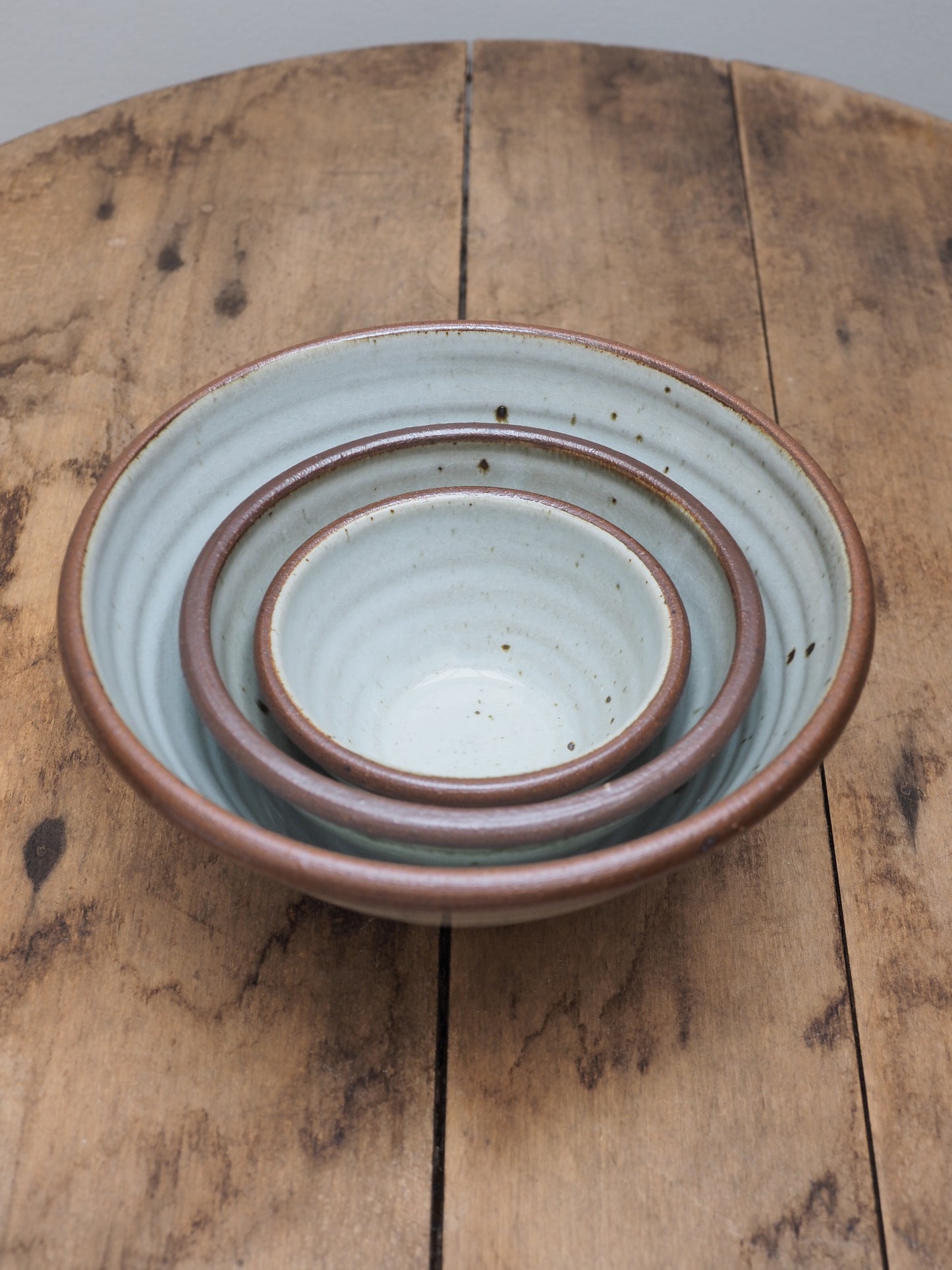 A set of three hand-thrown Leach Pottery stoneware bowls in three different sizes displayed nesting within each other on a wooden surface.