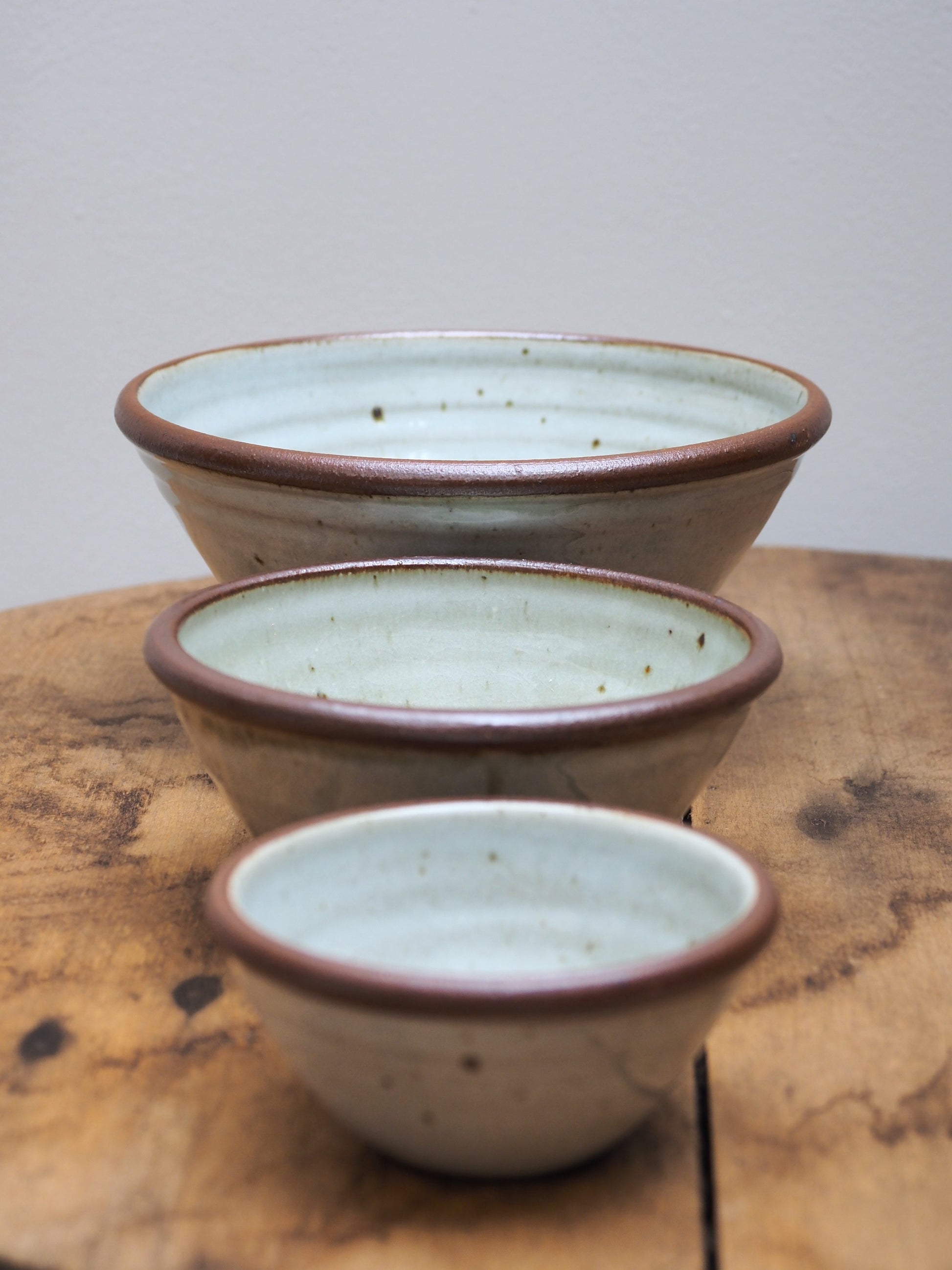 A set of three hand-thrown Leach Pottery stoneware bowls in three different sizes displayed one infront of the other on a wooden surface.