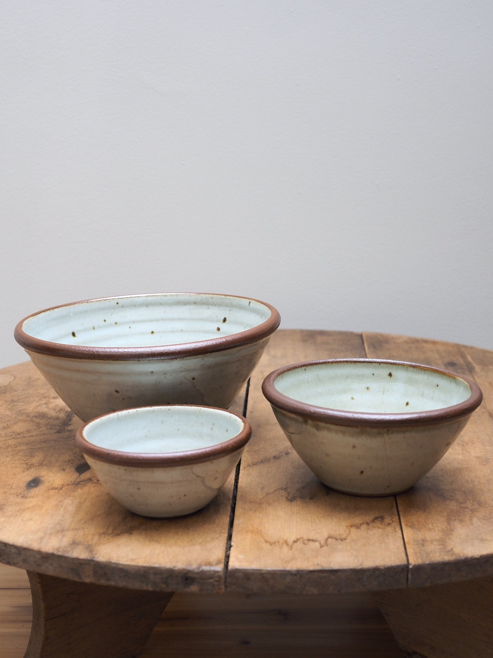 A set of three hand-thrown Leach Pottery stoneware bowls in three different sizes displayed next to each other on a wooden surface.