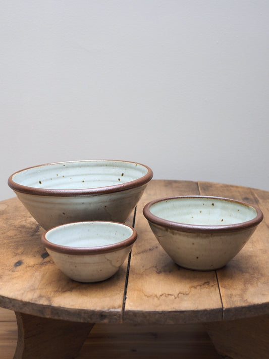 A set of three hand-thrown Leach Pottery stoneware bowls in three different sizes displayed next to each other on a wooden surface.