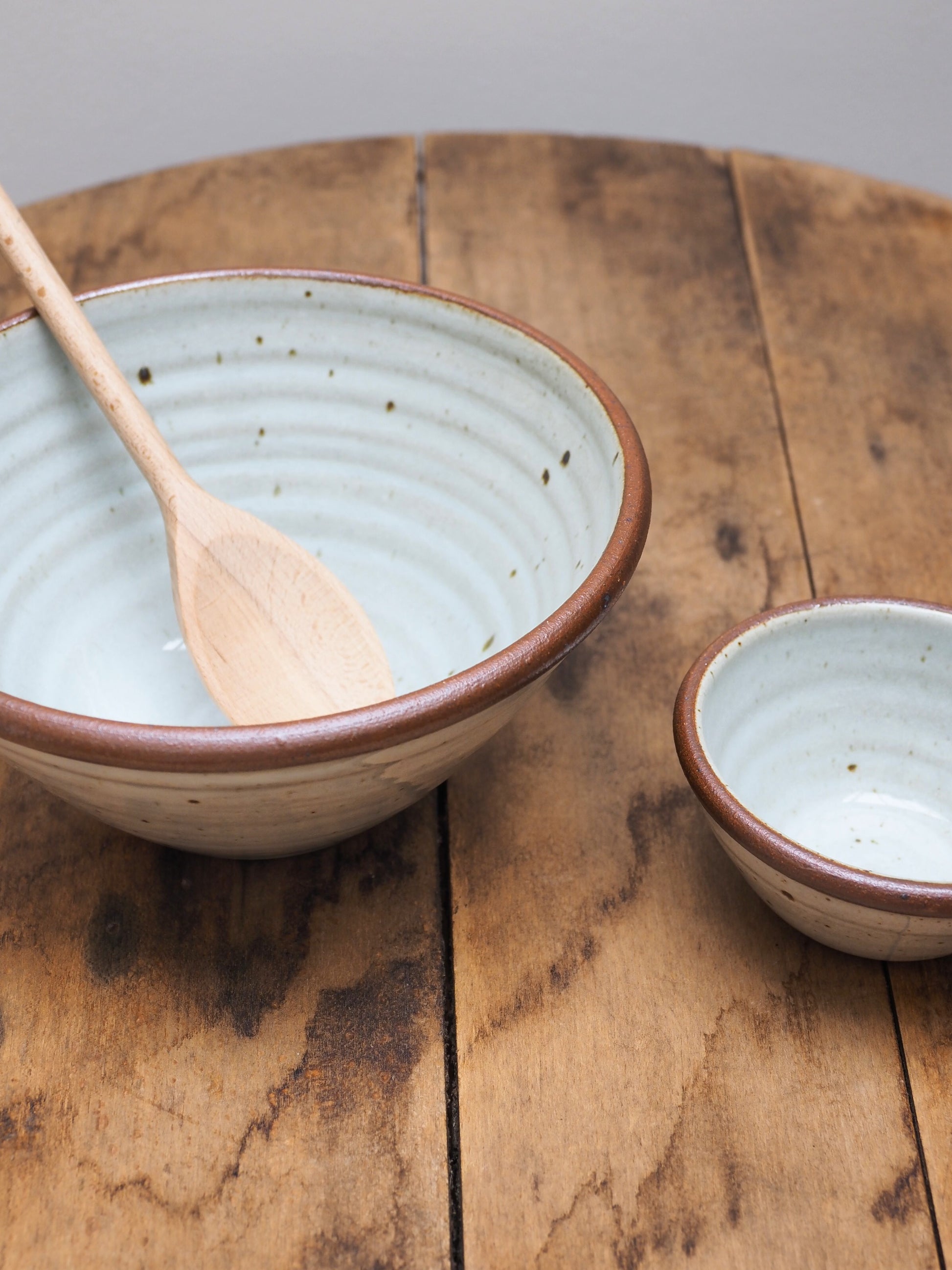 Close up of two hand-thrown Leach Pottery stoneware bowls displayed next to each other on a wooden surface, the larger bowl has a wooden spoon sticking out of it.