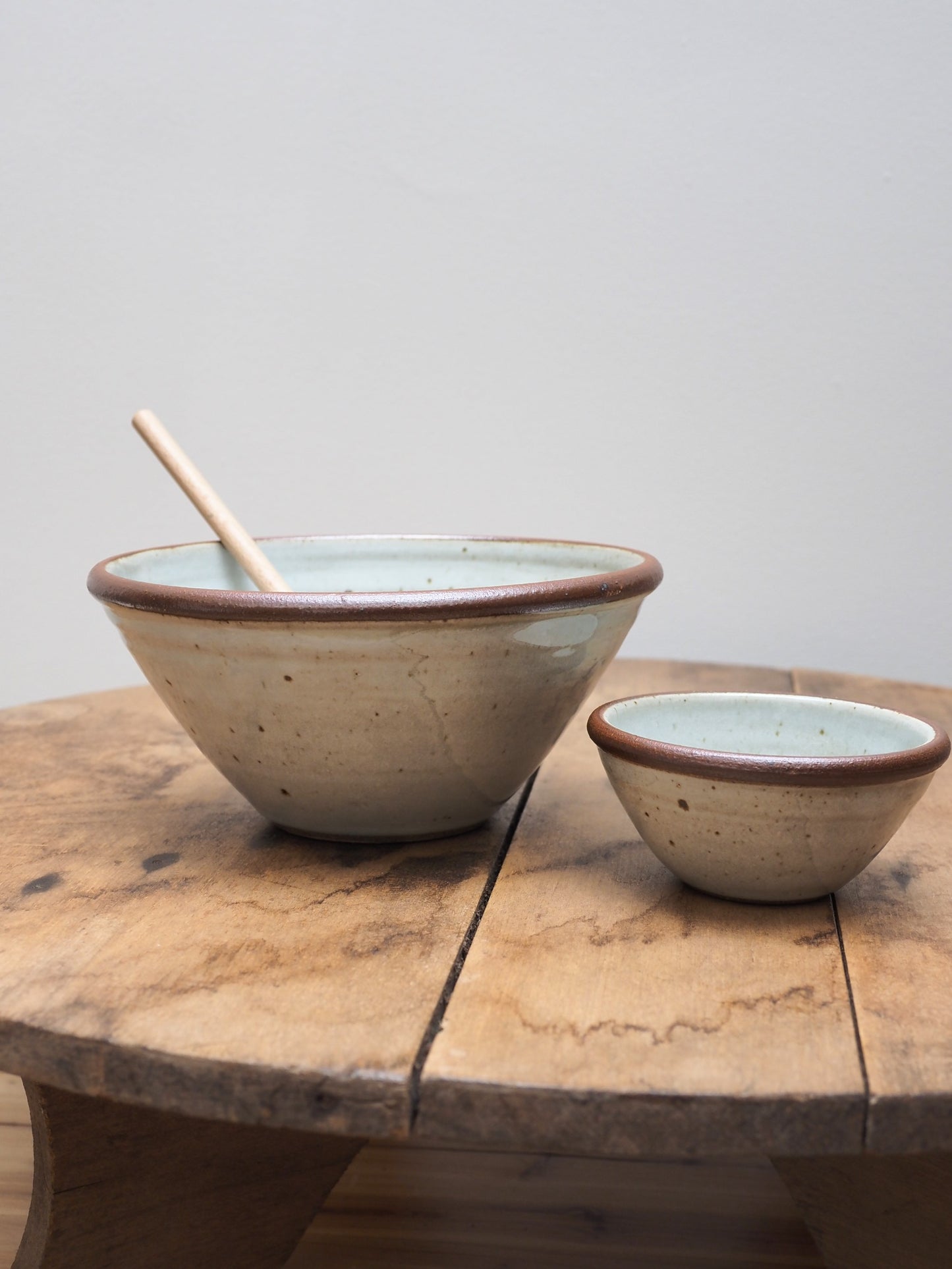 Two hand-thrown Leach Pottery stoneware bowls displayed next to each other on a wooden surface, the larger bowl has a wooden spoon sticking out of it.