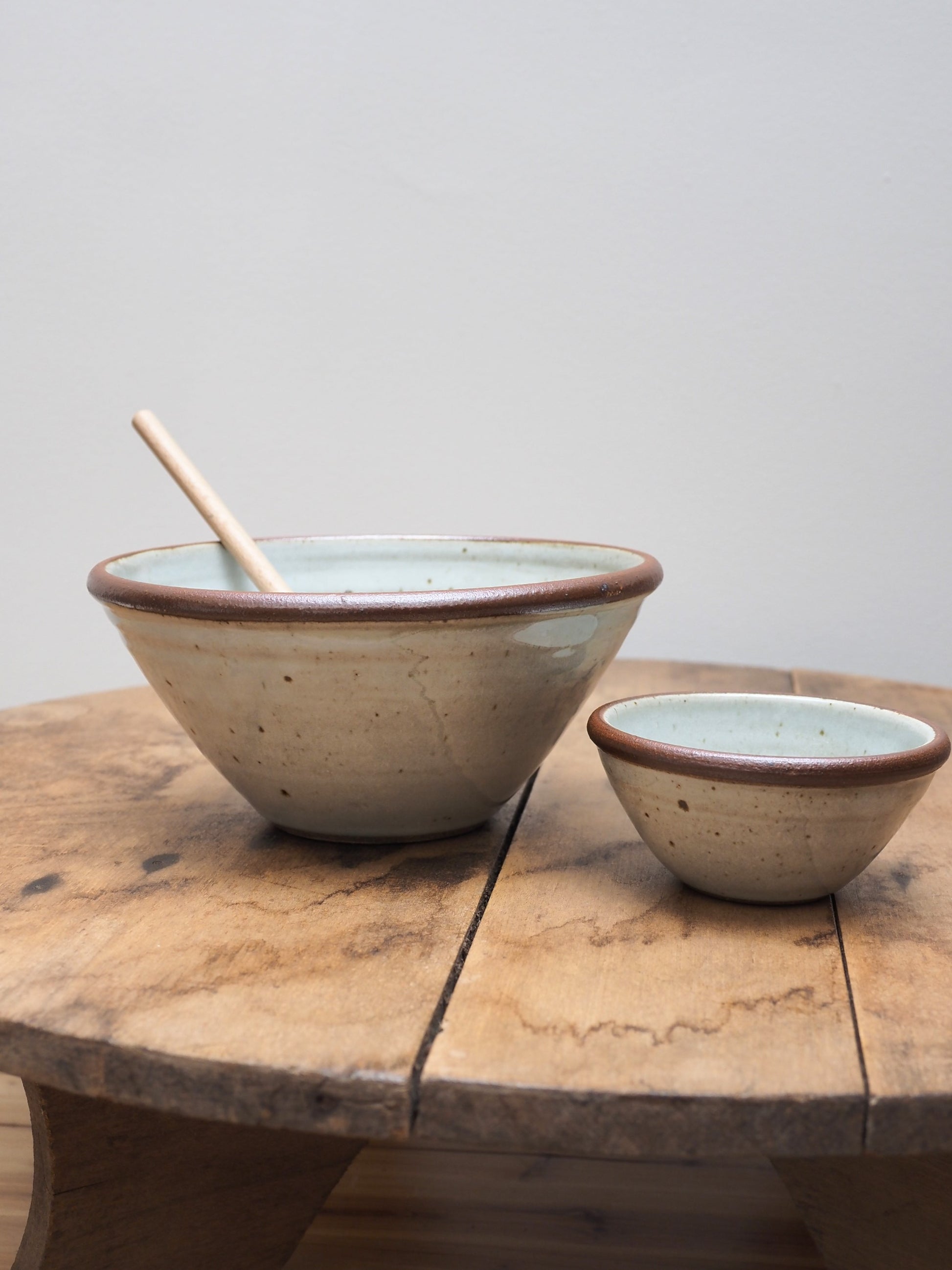 Two hand-thrown Leach Pottery stoneware bowls displayed next to each other on a wooden surface, the larger bowl has a wooden spoon sticking out of it.