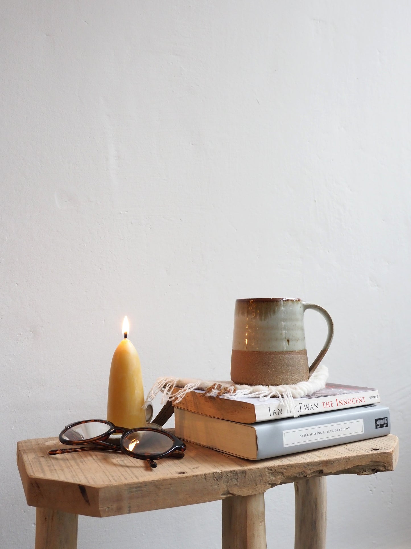 A Leach Pottery stoneware mug is on shown on top of a handmade woven coaster on top of a stack of books next to a lit candle and pair of glasses on a wooden table with a white background.