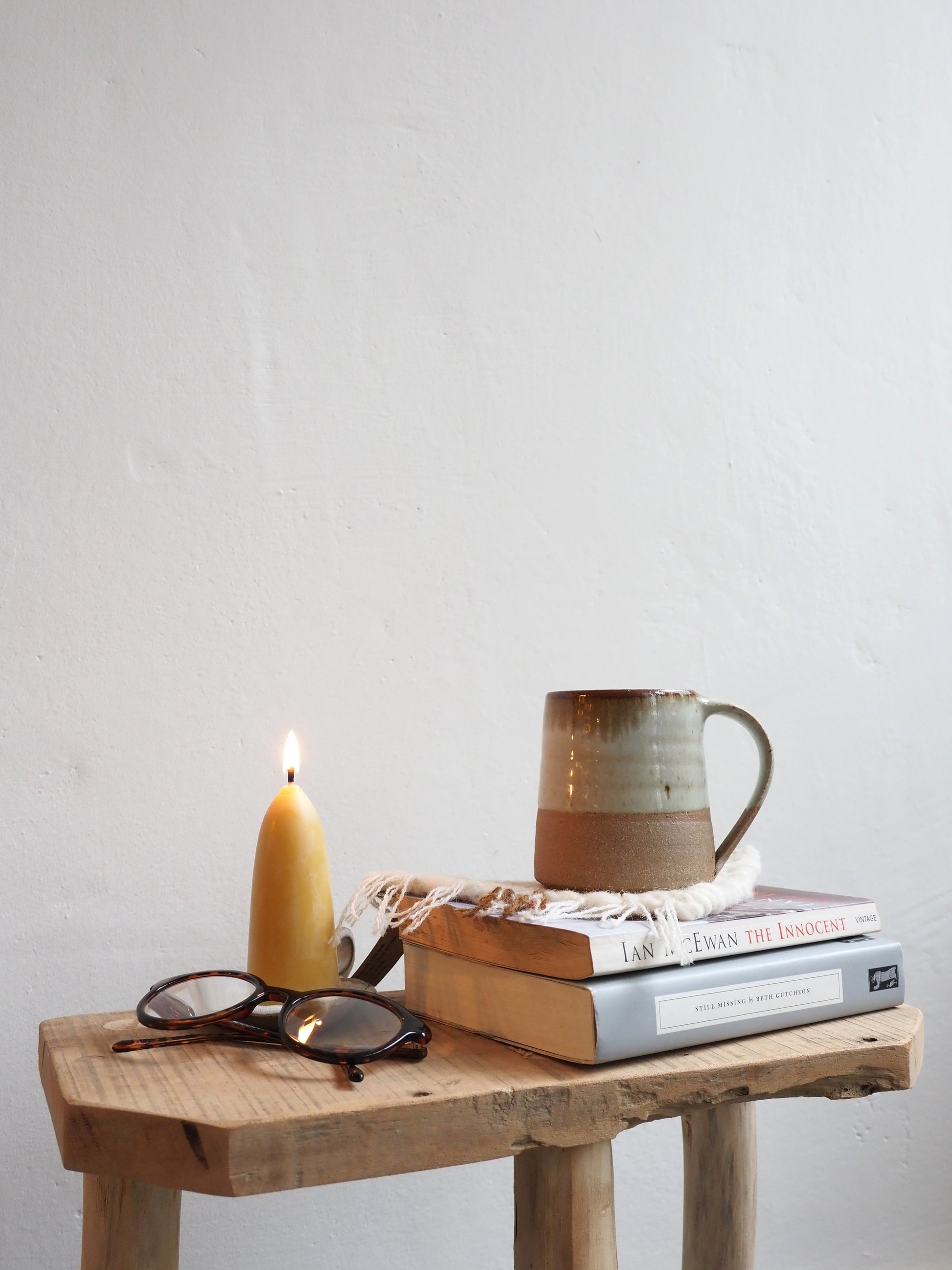 A Leach Pottery stoneware mug is on shown on top of a handmade woven coaster on top of a stack of books next to a lit candle and pair of glasses on a wooden table with a white background.