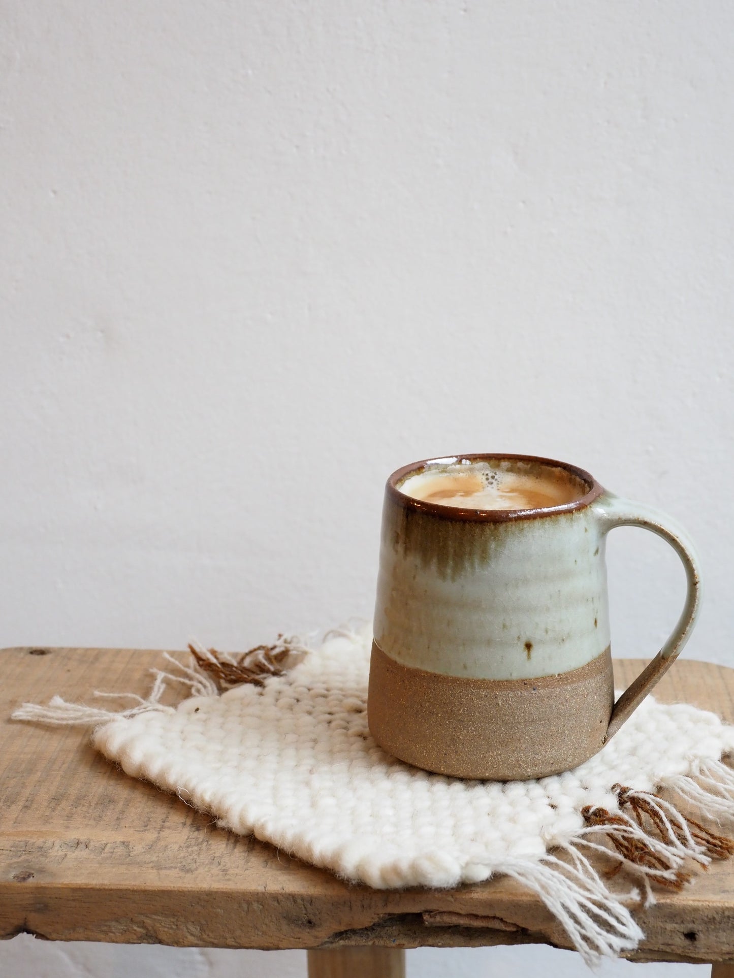 A Leach Pottery stoneware mug placed on handmade woven coaster on a wooden table agaisnt a white background.