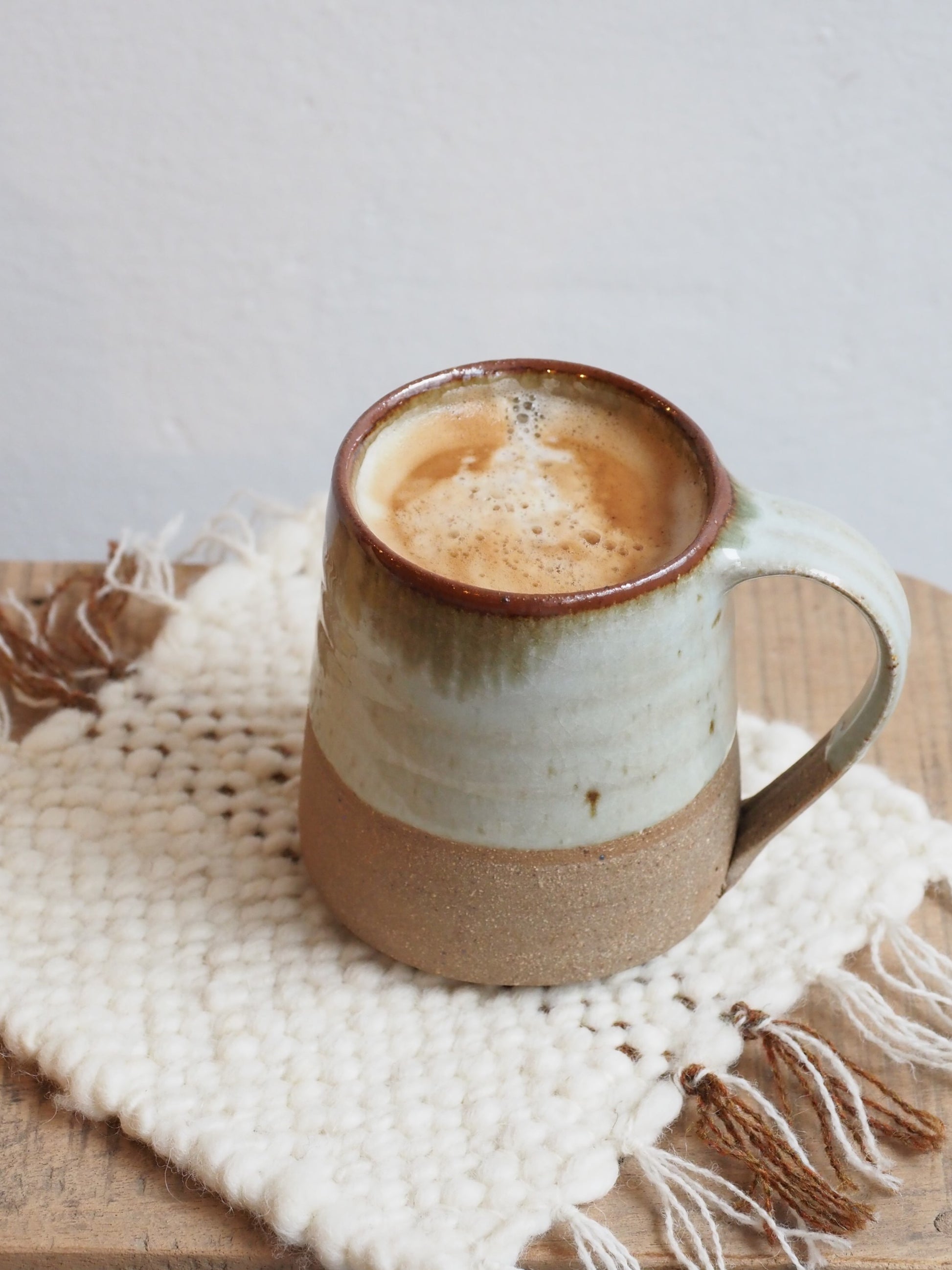 A Leach Pottery stoneware mug placed on handmade woven coaster on a wooden table agaisnt a white background.