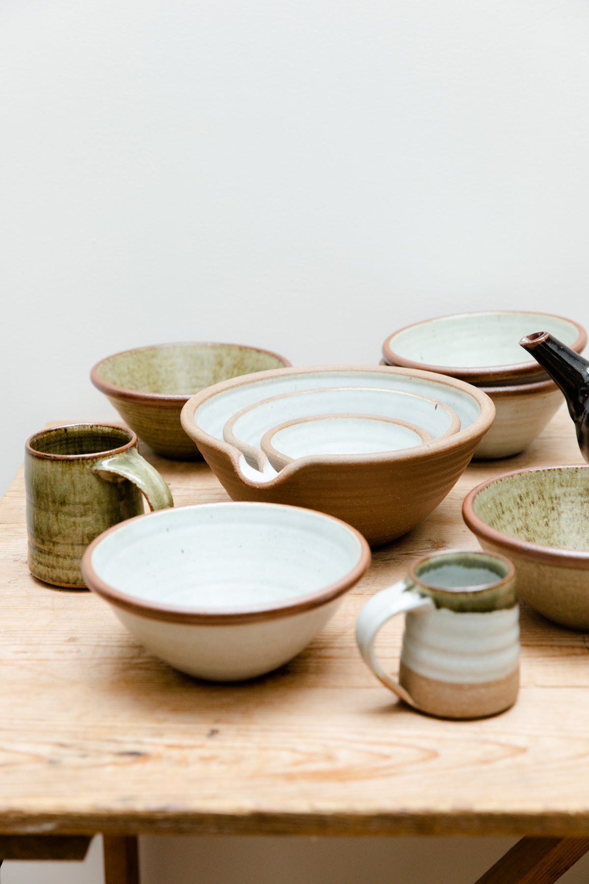 A collection of Leach Pottery assembled on a wooden table. Features a teapot, nesting mixing bowls, handthrown bowls and mugs.