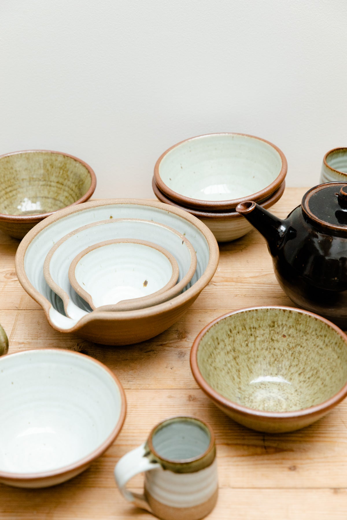 A collection of Leach Pottery assembled on a wooden table. Features a teapot, nesting mixing bowls, handthrown bowls and mugs.