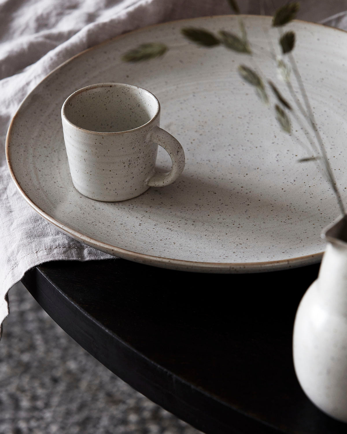 A white stoneware espresso cup placed on a matching stoneware plate, with a hint of greenery in the background.