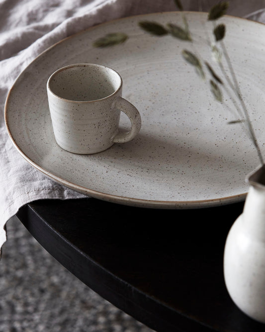 A white stoneware espresso cup placed on a matching stoneware plate, with a hint of greenery in the background.