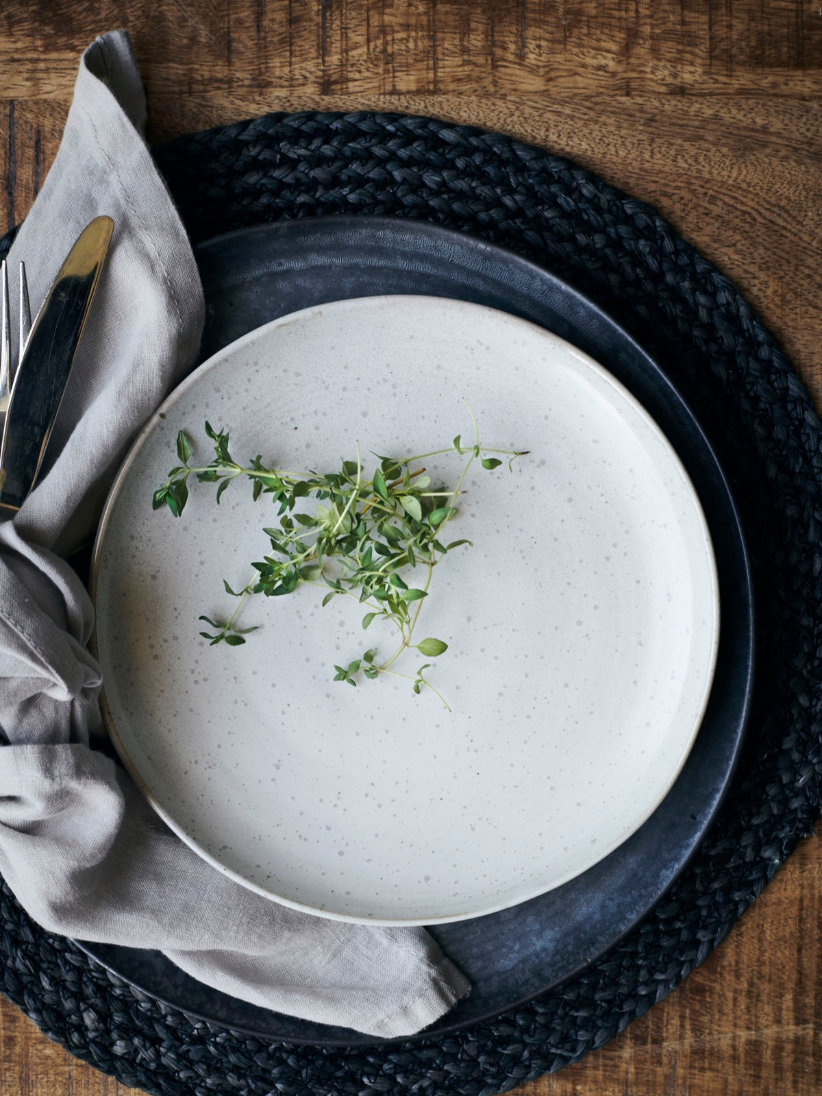 A stoneware lunch plate with a speckled white glaze, accompanied by a grey napkin and a dark grey placemat, set on a wooden table with a small amount of herbs placed on the plate.