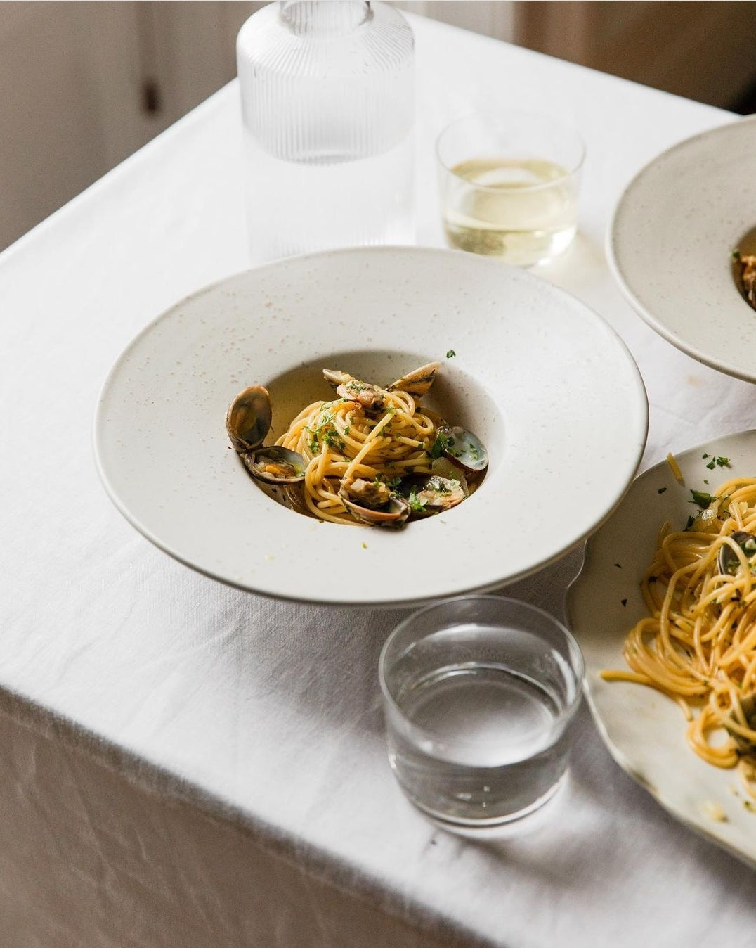 A white stoneware soup bowl with grey speckles, displayed on a table with food inside it, alongside a glass and another dish.