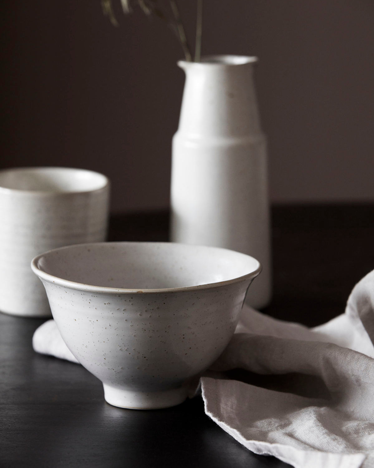 A white stoneware bowl with grey speckles on a table with a napkin and a vase in the background.