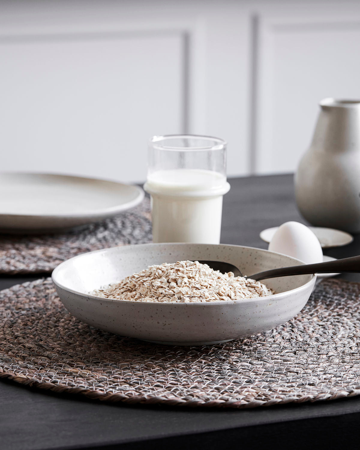 A wide stoneware bowl with a white and grey speckled glaze, containing cereal, placed on a table with a glass and a vase in the background.