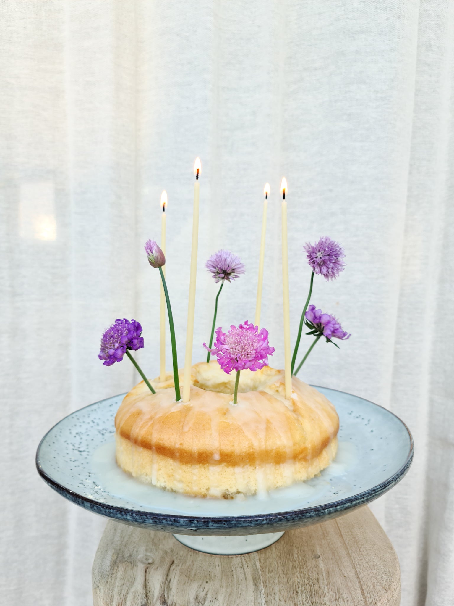Rustic cake stand holding a sponge cake with candles and flowers stuck into it.