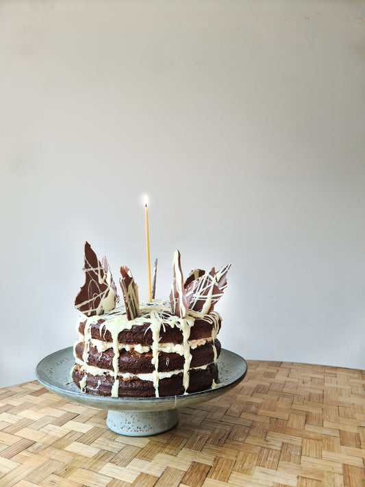 A chocolate cake with cream on a rustic cake stand with a single candle, displayed on a table.