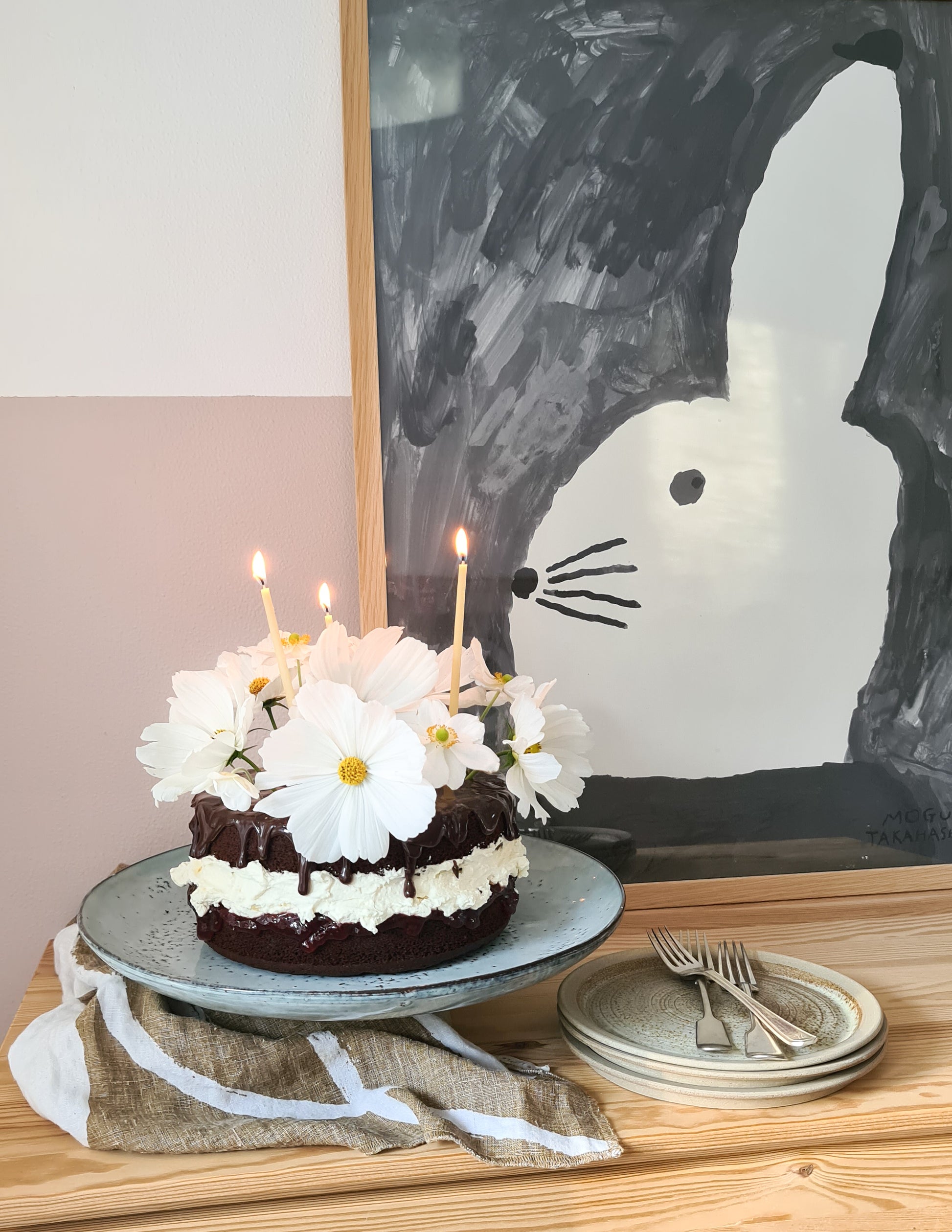 Rustic cake stand holding a chocolate cake with white flowers decorating it and lit beeswax candles next to a painting of a rabbit.