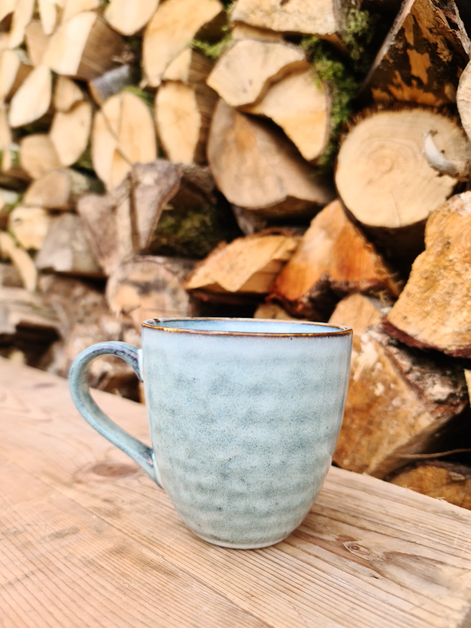 A blue/grey ceramic mug sitting in front of a log pile on a wooden table.