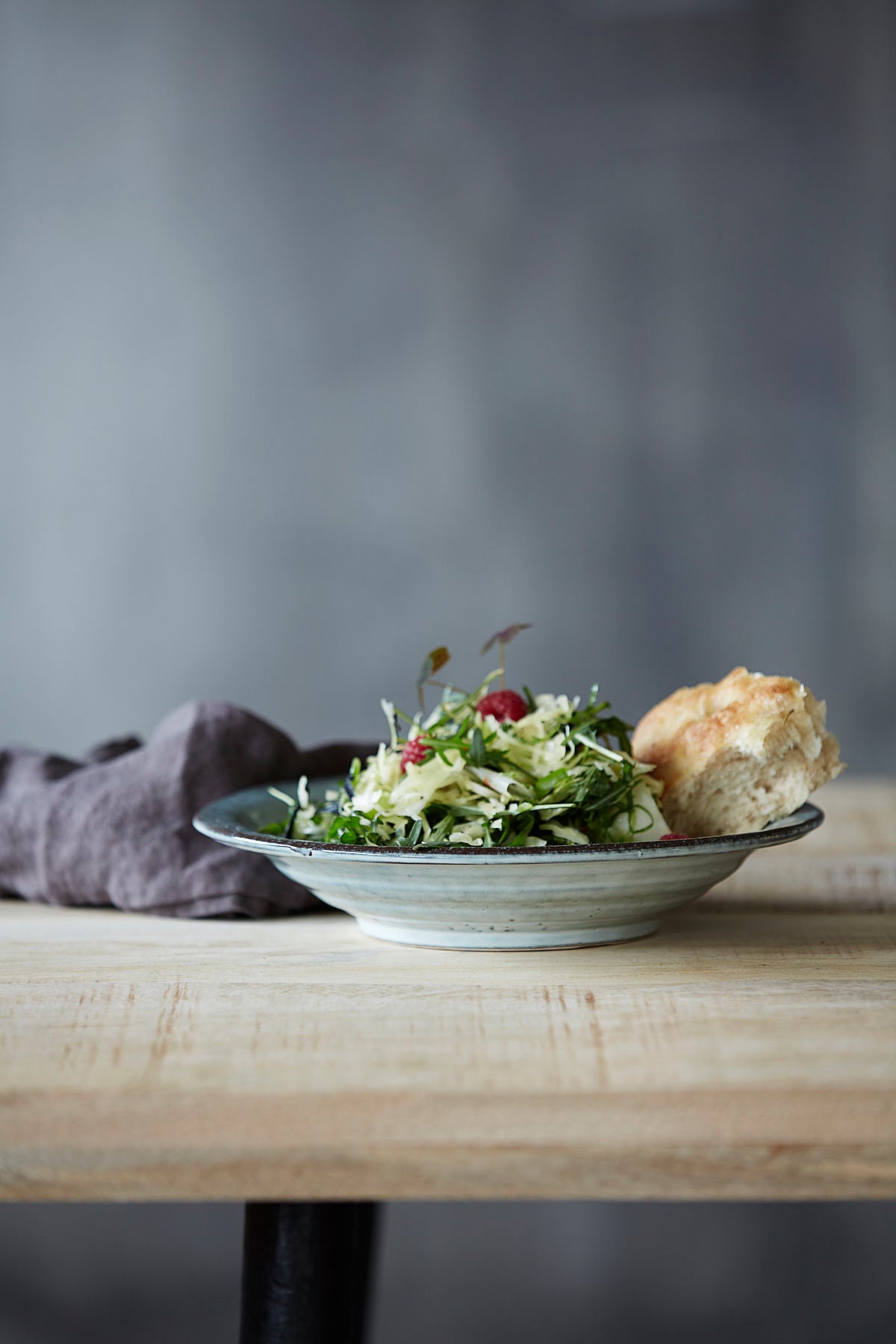A rustic grey blue pasta bowl set on a wooden table featuring a salad and bread.
