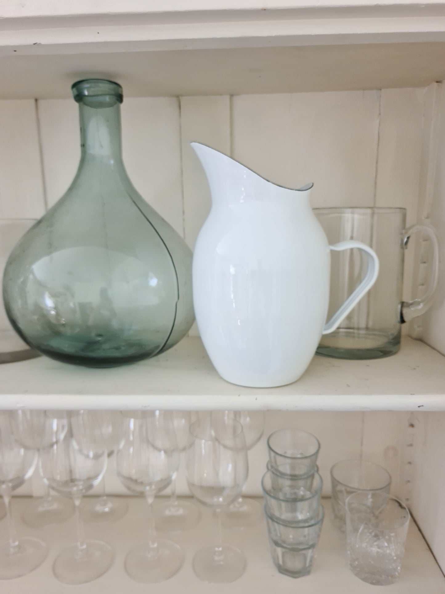 White enamel pitcher next to a glass vase and glasses on white shelving.