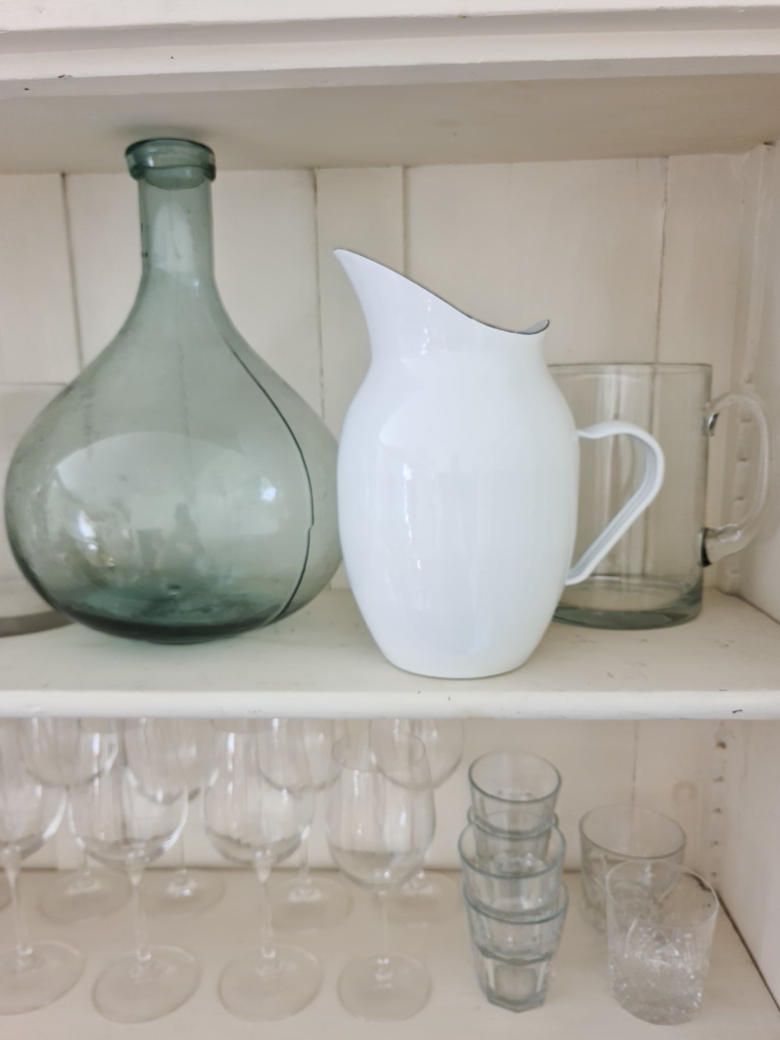 White enamel pitcher next to a glass vase and glasses on white shelving.