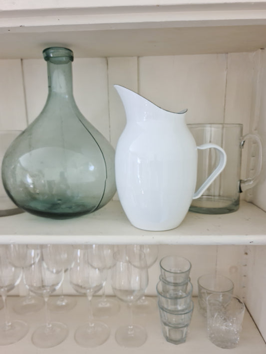 White enamel pitcher next to a glass vase and glasses on white shelving.