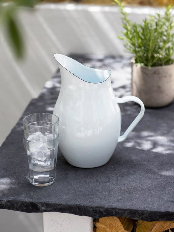 White enamel pitcher on a slate table next to two stacked glasses and a potted plant in the background.