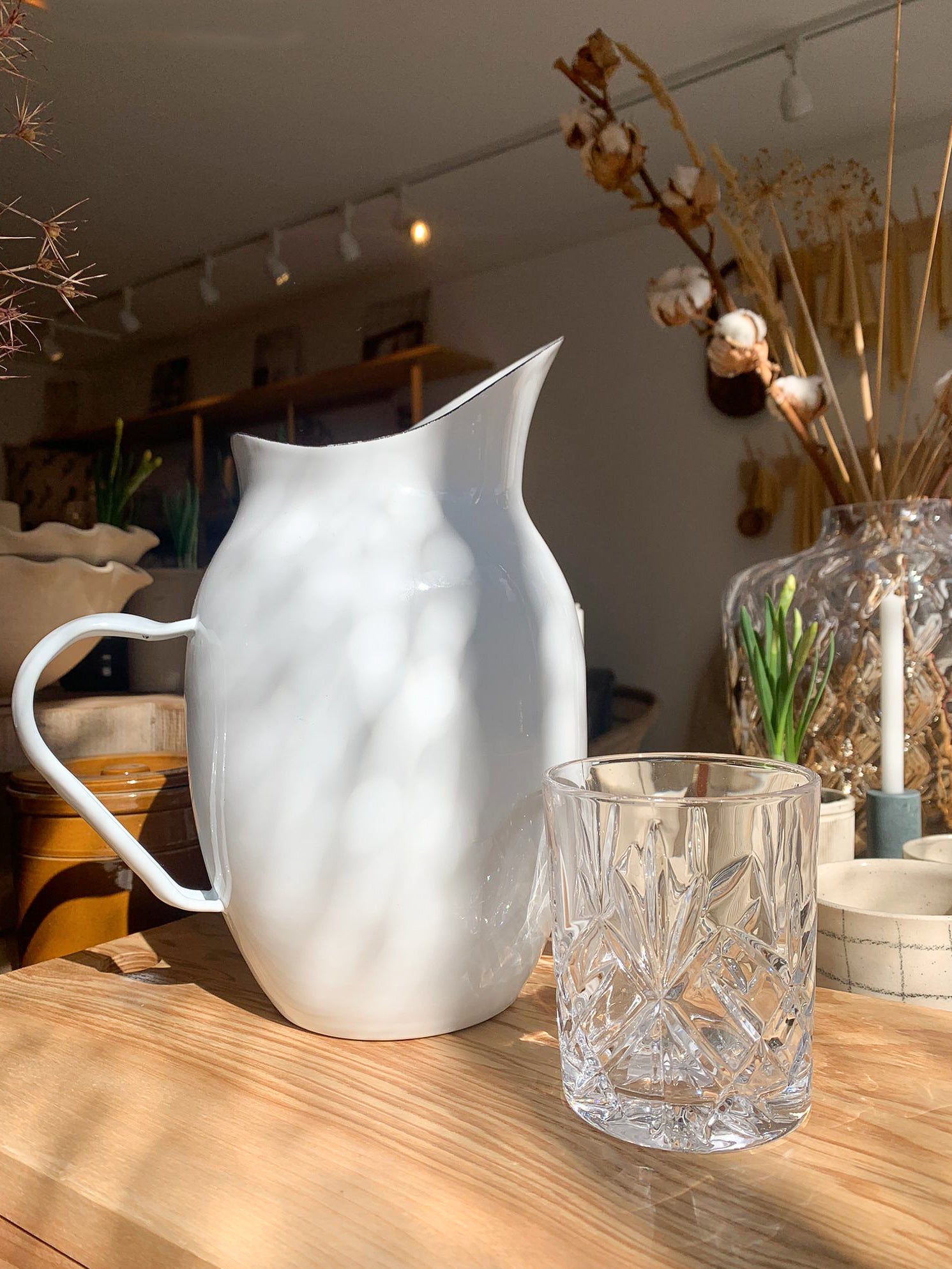 White enamel pitcher next to a scotch glass on a wooden board.
